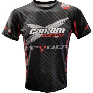 May include: Black short-sleeved t-shirt with a carbon fiber pattern and red accents. The shirt features the "Can-Am Team Spyder" logo in white and red across the chest, with the BRP logo on the upper right shoulder.