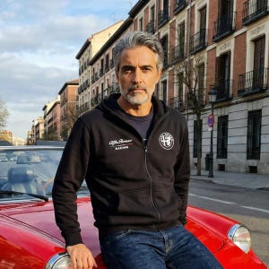 Może przedstawiać: Czarna bluza z kapturem z logo Alfa Romeo Racing na piersi i okrągłą odznaką na rękawie. Osoba ma na sobie niebieskie dżinsy i opiera się o czerwony zabytkowy samochód.