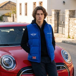 May include: A bright blue puffer vest with the Mini Cooper logo and a Union Jack patch. The vest is worn over a black hooded sweatshirt. A red Mini Cooper car is in the foreground.