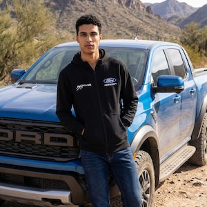 May include: A black zip-up hoodie featuring the Ford Ranger logo and Raptor text in white. The hoodie is paired with blue jeans. A blue Ford Ranger pickup truck is in the background, with a desert landscape.