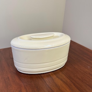 May include: An oval-shaped, cream-colored ceramic butter dish with a matching lid. The base of the dish features three horizontal embossed lines circling the exterior, and the lid has a smooth, slightly raised handle on top.