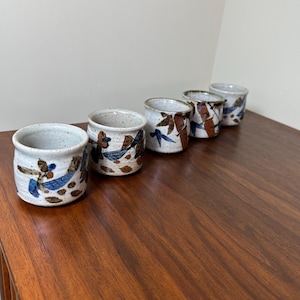 May include: A set of five small ceramic cups with a speckled off-white glaze. Each cup features a hand-painted design of blue leaves and brown stems or branches wrapping around the exterior, with a smooth, rounded rim and a cylindrical shape.