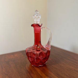 Pode incluir: Uma garrafa de vidro vermelha com uma rolha de vidro transparente e uma alça. A garrafa tem uma superfície texturizada e com bolhas.