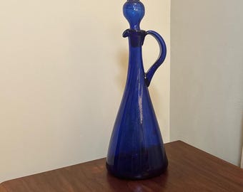 Può includere: Una caraffa in vetro blu cobalto con un beccuccio svasato e un manico. La caraffa ha una superficie testurizzata e si trova su un tavolo di legno.
