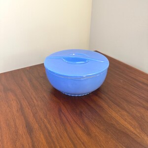 May include: A light blue, round container with a matching lid. The container is sitting on a dark brown wooden surface. The lid has a small handle in the center.