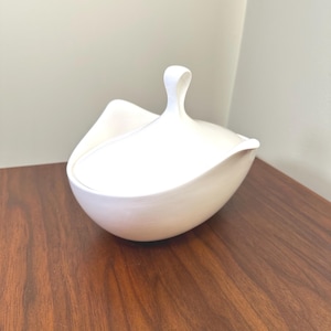 May include: A white ceramic lidded bowl with a unique, organic shape. The lid features a curved handle. The bowl is set on a dark wood surface, creating a contrast with the bright white ceramic.
