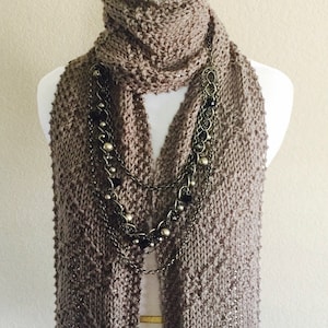 May include: A brown knitted scarf with a chain necklace featuring silver and black beads. The scarf is displayed on a white mannequin.