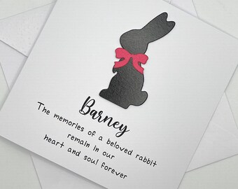 May include: A square white greeting card featuring a black silhouette of a rabbit with a pink bow around its neck. The text reads: Barney. The memories of a beloved rabbit remain in our heart and soul forever. The card is placed on a white envelope.