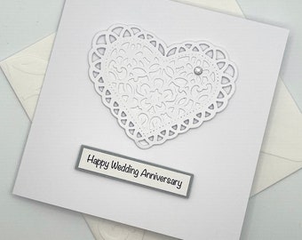 May include: A square white greeting card featuring a die-cut white paper heart with an intricate lace-like pattern and a single small clear rhinestone accent. Below the heart, a rectangular label displays the text Happy Wedding Anniversary.