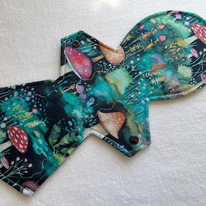 Può includere: Assorbente mestruale in tessuto riutilizzabile con un fantasioso design a tema foresta. L'assorbente presenta uno sfondo verde acqua scuro con illustrazioni di funghi, piante e fiori nei toni del rosso, rosa e marrone. Ha bottoni automatici.