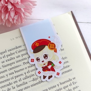 Puede incluir: Un marcapáginas con una ilustración de dibujos animados de una niña con cabello rubio que lleva un sombrero rojo y un vestido rojo. Ella sostiene un pequeño animal blanco en sus brazos. El marcapáginas está sobre un fondo azul claro con flores blancas.