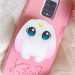 Puede incluir: Una funda de teléfono rosa con un agarre de teléfono de conejo blanco. El conejo tiene grandes ojos azules y una luna creciente en la cabeza. La funda del teléfono tiene la palabra "alien" escrita en ella.