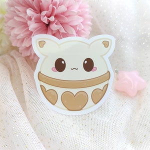 Puede incluir: Una pegatina blanca con una ilustración de dibujos animados de un personaje lindo, redondo, marrón y blanco con ojos grandes y un patrón en forma de corazón. El personaje tiene un patrón en forma de corazón marrón claro alrededor del medio de su cuerpo.