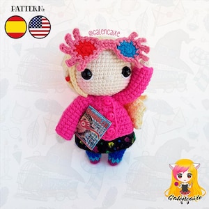 May include: A crocheted doll wearing pink glasses, a pink sweater, a black and blue skirt, and holding a book. The doll has blonde hair and brown eyes. The book has the title "The Monster Book of Monsters" on the cover.