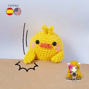 Op de afbeelding: Een geel gehaakt kuiken speelgoed met zwarte ogen, een oranje snavel en roze wangen. Het kuiken valt en heeft een verraste uitdrukking. Het speelgoed ligt op een bruin oppervlak met een zwart-witte comic-stijl geluidseffect.