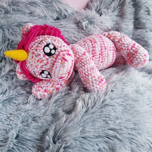 Può includere: Un giocattolo di unicorno all'uncinetto rosa e bianco con corno giallo sdraiato su una morbida coperta grigia.