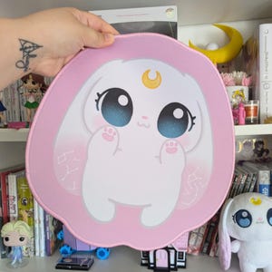 May include: A pink and white mousepad featuring a cartoon bunny with large blue eyes and a golden crescent moon. The bunny is set against a pink background with Japanese text. The mousepad is round and has a stitched edge.