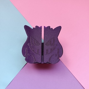 Puede incluir: Una figura de Pokémon fantasma impresa en 3D de color morado con una sonrisa amenazante. La figura está dividida por la mitad, creando un diseño simétrico.