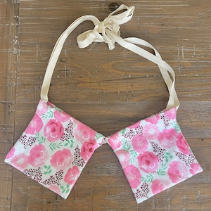 Puede incluir: Un par de bolsas de tela floral rosa con correas blancas. Las bolsas están conectadas por las correas y se pueden llevar alrededor del cuello.