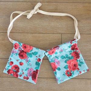 Puede incluir: Dos bolsas de tela con estampado floral y una correa de tela crema. Las bolsas están atadas juntas en la parte superior y tienen un fondo azul con rosas rojas y rosas.