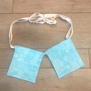May include: A light blue fabric item with two square pockets, connected by a white ribbon. The pockets have a floral pattern in white. The ribbon is tied in a bow at the top.
