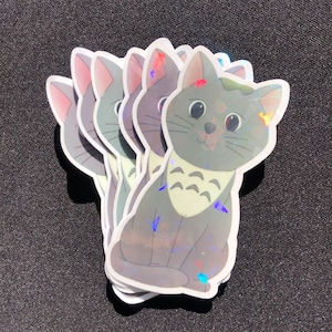 Może przedstawiać: Stos pięciu tęczowych szarych naklejek z kotem w białym kołnierzyku i wzorem przypominającym Totoro. Naklejki są holograficzne i mają błyszczące wykończenie.