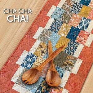 May include: A red and white quilted table runner with a patchwork design featuring various floral and geometric patterns. Two wooden serving spoons are placed on top of the runner. The text "CHA CHA CHA!" is printed in black at the top of the runner.