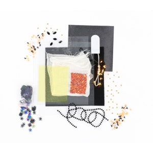 May include: A Halloween craft kit with a variety of materials, including black and gold glitter, black beads, orange and red sequins, black and white confetti, and a small bag of orange and red rhinestones. The kit also includes a white tulle bag, a black felt sheet, and a string of white fairy lights.