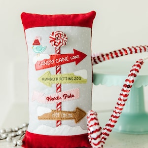 May include: A gray and white pillow with red trim features a red, white, and green candy cane with a red and white bow.  The pillow has four signs with the text "Candy Cane Lane", "Reindeer Petting Zoo", "North Pole", and "Hot Cocoa".