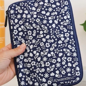 May include: A navy blue zippered case with a white floral pattern. The case is rectangular with rounded corners and a black zipper. The brand name "KIMBERBELL" is printed on the bottom right corner.