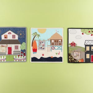 May include: Three decorative fabric wall hangings depicting houses and scenes. One shows a house with fireworks and an American flag. Another features a beach scene with a surfboard. The third depicts a house with a tree and pumpkins.
