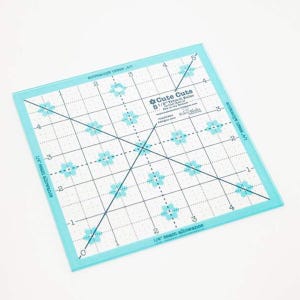 May include: A clear, square quilting ruler with a light blue border. The ruler features a grid pattern with flower designs and measurements in inches. Text on the ruler includes "Cute Cuts" and "5 1/2" Trim-it Ruler".