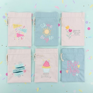 Puede incluir: Seis pequeñas bolsas con cordón en colores pastel. Cada bolsa presenta diseños y texto bordados, incluyendo "You Did It", un sol con "Shine Bright", flores, "Friends are the family we pick for ourselves", "Congrats!", una magdalena con "Happy Birthday" y "Big Hugs".