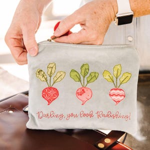 Puede incluir: Un estuche gris claro con cremallera y bordados de rábanos, con el texto "Darling, you look Radishing!". El estuche presenta tres diseños de rábanos en rojo, rosa y blanco con hojas verdes. El tirador de la cremallera es rojo.