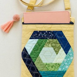 Puede incluir: Bolso bandolera acolchado amarillo con un diseño de patchwork geométrico en tonos azules, verdes y blancos. El bolso tiene una borla rosa y una correa a juego. Una tableta rosa es visible dentro.