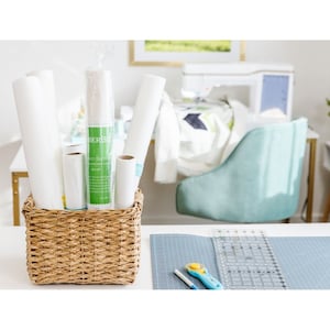 May include: A wicker basket filled with rolls of white fabric, including a roll labeled "Perfect Barrier" with green text. The basket is on a white table with a gray cutting mat and a rotary cutter.
