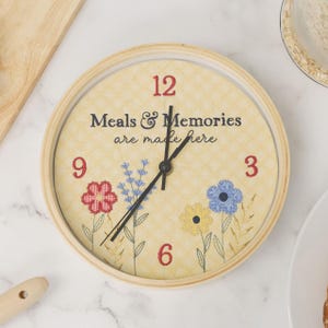 May include: A round wall clock with a light yellow checkered background. The clock face features the text "Meals & Memories are made here" in black, red numbers 3, 6, 9, and 12, and embroidered floral designs in red, blue, and yellow.