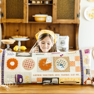 May include: A decorative rectangular pillow with pie-themed designs. The pillow features various pie slices, a mixer, and the text "GATHER TOGETHER, GIVE THANKS, EAT PIE." The color palette includes orange, purple, and cream tones, with a patchwork design.