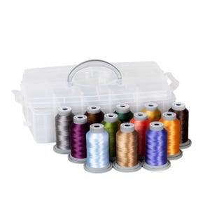 Könnte beinhalten: Eine transparente Kunststoff-Organizer-Box mit Griff, gefüllt mit Spulen aus farbigem Stickgarn. Die Garnfarben umfassen Grau, Lila, Hellblau, Grün, Gold, Rot, Blau und Orange. Die Spulen sind vor dem Organizer angeordnet.