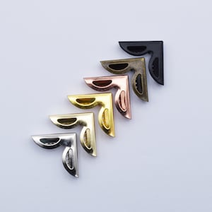 May include: Five metal corner protectors in different colours: black, bronze, copper, gold, and silver. The corner protectors are arranged in a row, with the black one at the top and the silver one at the bottom.