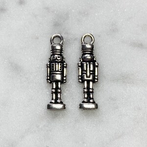 May include: Two silver-toned nutcracker charms with a loop at the top for hanging. The charms are detailed with a hat, face, and body.