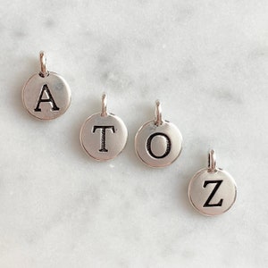 May include: Four small, round, silver charms, each bearing a capital letter: A, T, O, and Z. Each charm has a small loop for attaching to jewellery. The letters are in a bold, sans-serif font. The charms are displayed on a light-colored surface.