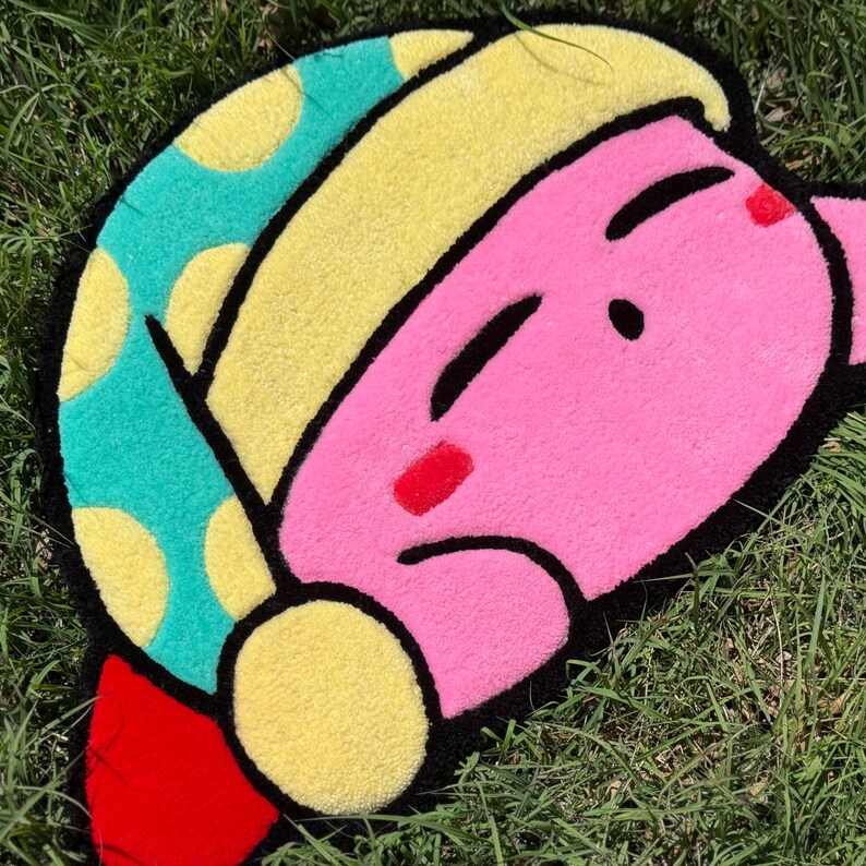 May include: A pink and yellow plush rug shaped like a cartoon character with a sleepy expression. The character has a blue and yellow hat with white polka dots.