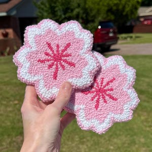 May include: Two pink and white crocheted coasters shaped like cherry blossoms. The coasters have a pink flower design in the centre.