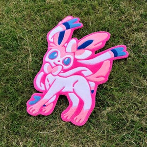 May include: A pink and white plush rug shaped like a cartoon character with blue details. The rug is on a green grassy surface. The character has large blue eyes and a smiling expression.