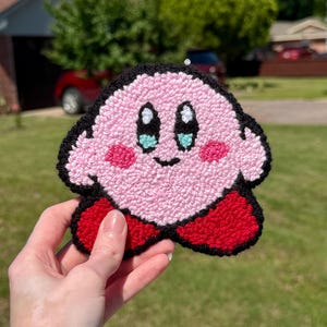 May include: Pink and red felt character with black outlines and blue eyes. The character is a video game character named Kirby.
