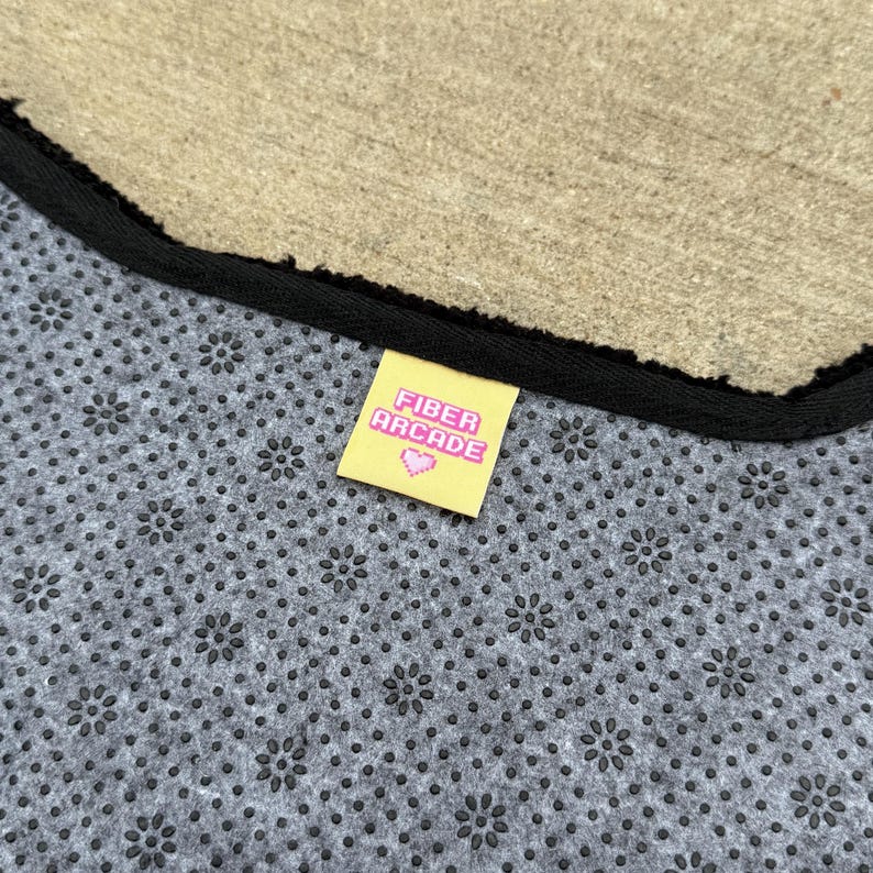 Pu&ograve; includere: Un tappeto grigio con un motivo floreale e un bordo nero. Un'etichetta gialla con la scritta "FIBER ARCADE" e un cuore rosa &egrave; attaccata al tappeto.