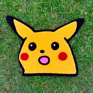 May include: Yellow character rug with black outlines and details. The rug features black eyes, red cheeks, and a pink mouth. The rug is placed on a green grassy surface.