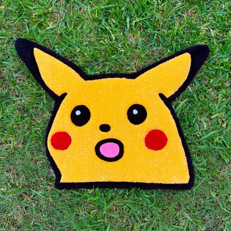 May include: Yellow character rug with black outlines and details. The rug features black eyes, red cheeks, and a pink mouth. The rug is placed on a green grassy surface.