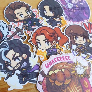 May include: A selection of cartoon character stickers in various colours. The stickers depict detailed characters with bright colours. Some stickers have text, including "WHEEEEEEE" and "VIOLE". The stickers are on a light brown surface.